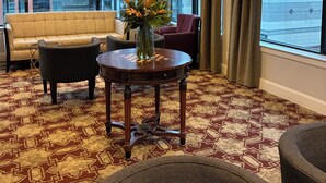 A hotel room with a large window, a chandelier, a sofa, a round table with a vase, and patterned carpet.