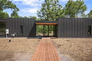 Ein modernes Gebäude mit einer Holzterrasse, die zu einem Glastürbereich führt, umgeben von Bäumen.