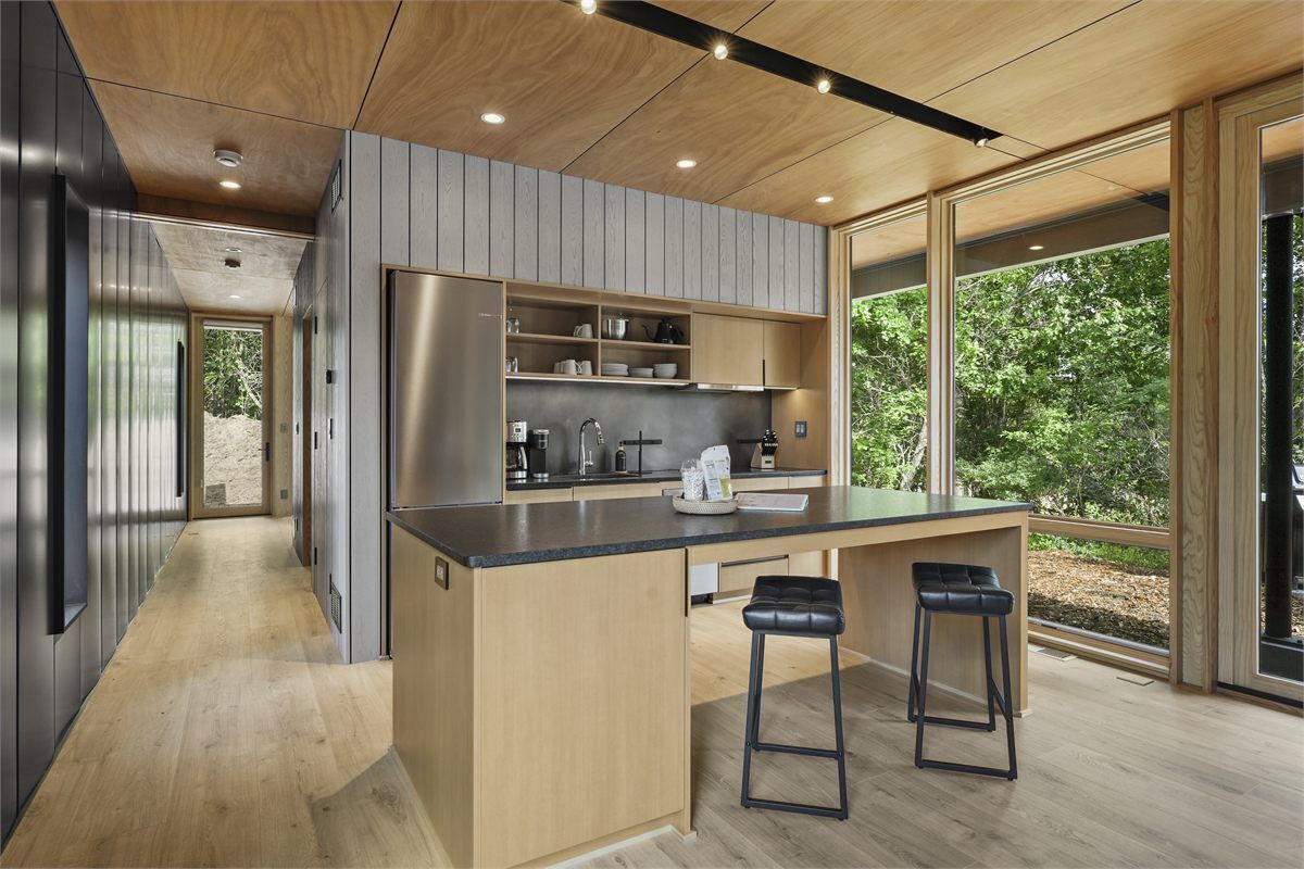 A modern kitchen with a central island, bar stools, and a view of greenery outside.
