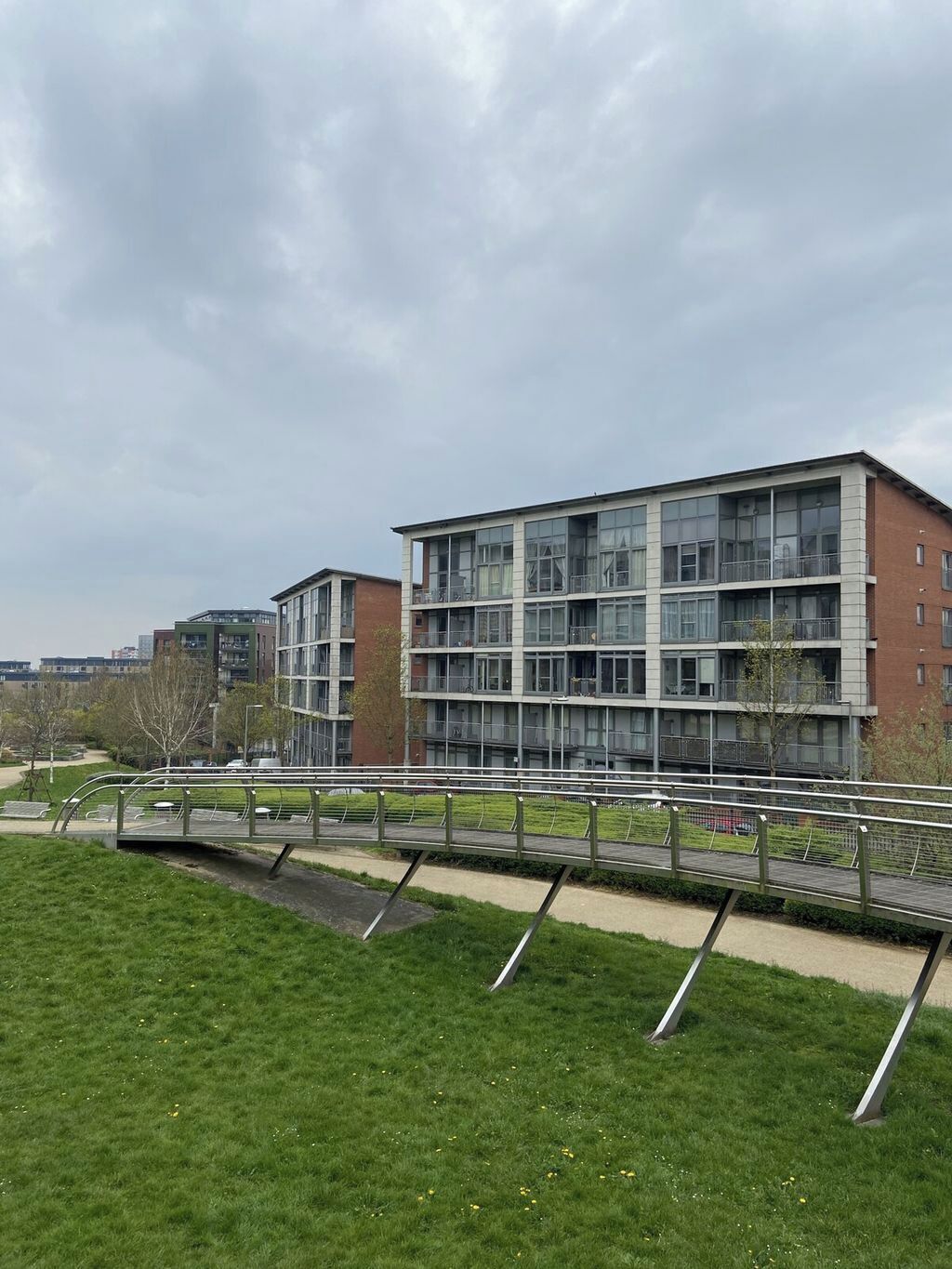 A modern multi-story building with large windows and balconies, surrounded by a grassy area and a pedestrian bridge.