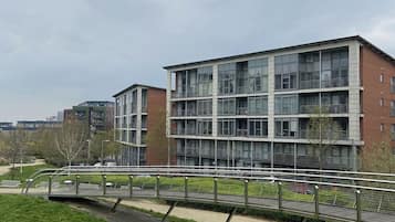 A modern multi-story building with large windows and balconies, surrounded by a grassy area and a pedestrian bridge.