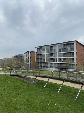 Un immeuble moderne à plusieurs étages, doté de grandes fenêtres et de balcons, entouré d’une zone verdoyante et d’un pont piétonnier.
