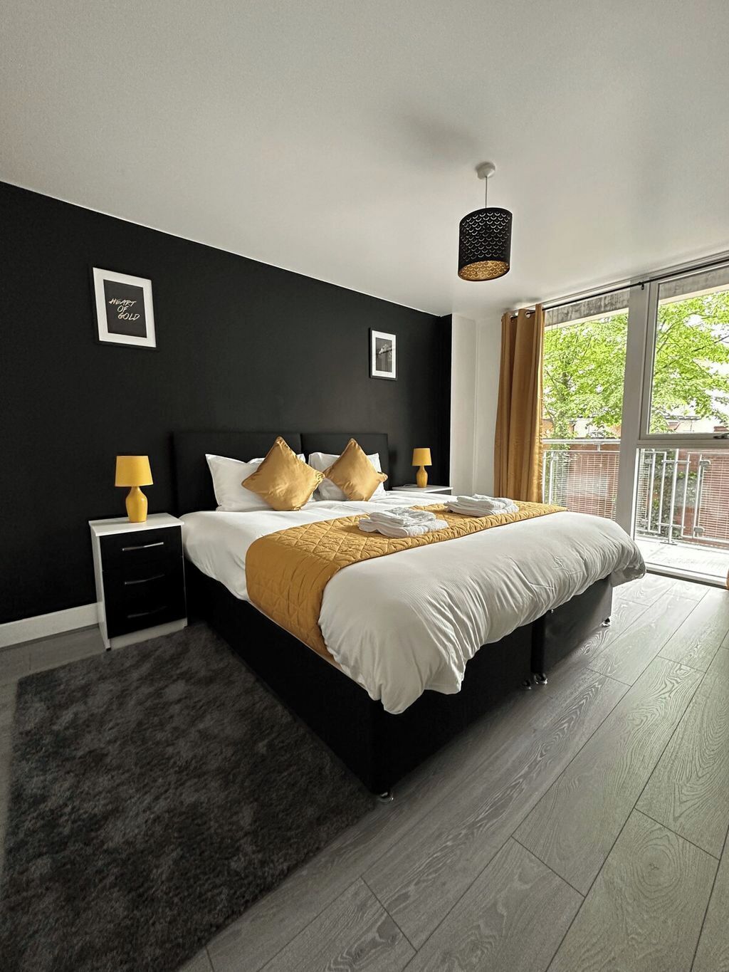 A modern bedroom with a black wall, a large bed with white and gold bedding, two bedside tables with lamps, and a view of greenery outside.
