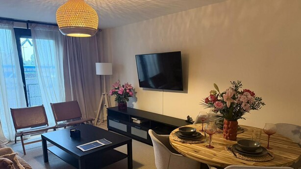 A modern hotel room with a dining table set for two, a flat-screen TV mounted on the wall, and a large window with curtains. - City Center Gem Steps from Albert Clock &Cathedral (Belfast)