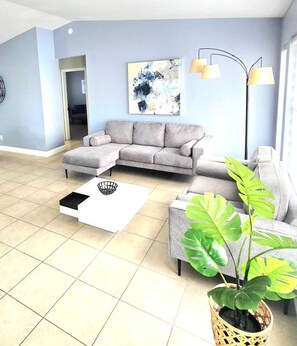 Ein Wohnzimmer mit Ecksofa, Couchtisch, Zimmerpflanze und Wandlampe.