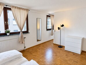Ein Hotelzimmer mit einem Bett, einem Nachttisch, einem Schminktisch, einem Spiegel, einer Stehlampe und zwei mit Vorhängen versehenen Fenstern.