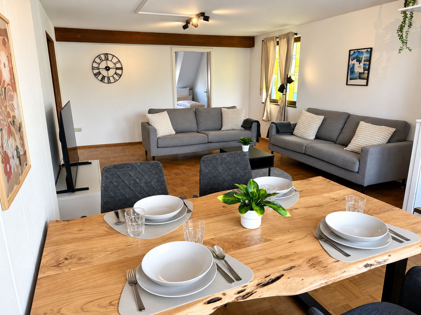 A modern living room with a dining area, grey sofas, a wooden dining table, and a clock on the wall.