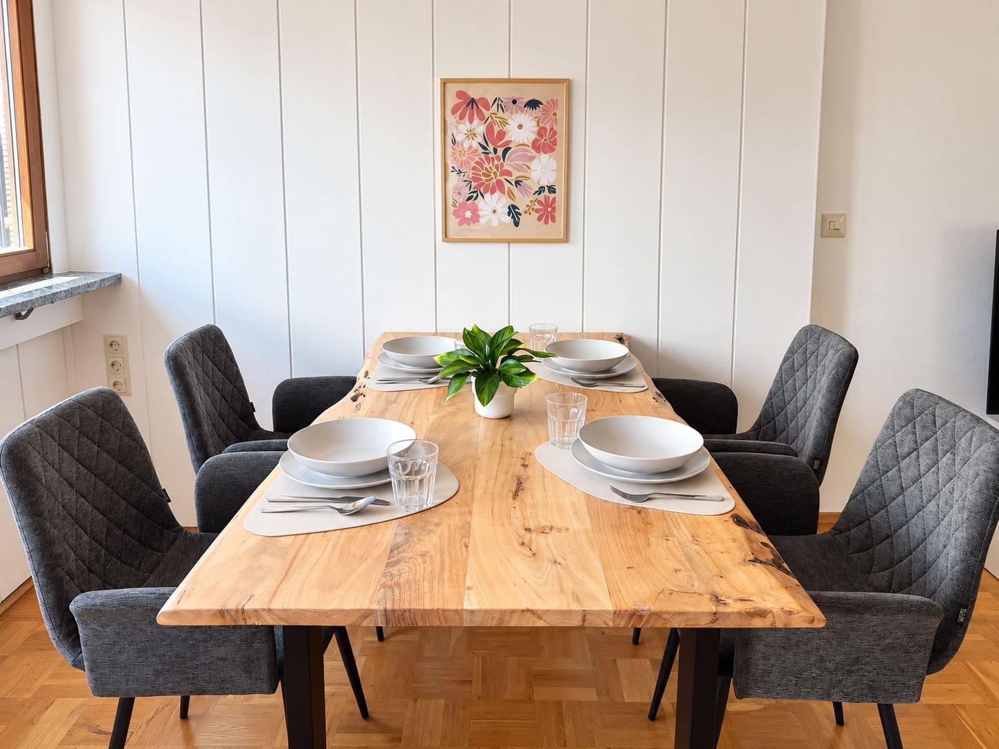 Et spisebord dækket med tallerkener, glas og bestik, omgivet af polstrede stole, med en potteplante på bordet.