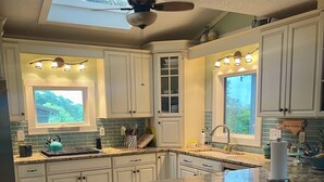 A kitchen with white cabinets, a skylight, a ceiling fan, and a view of greenery outside. - Fall for Canal Breeze: Private Lot on the Canal, Saltwater Pool; Dock; Beach! (Oak Island)