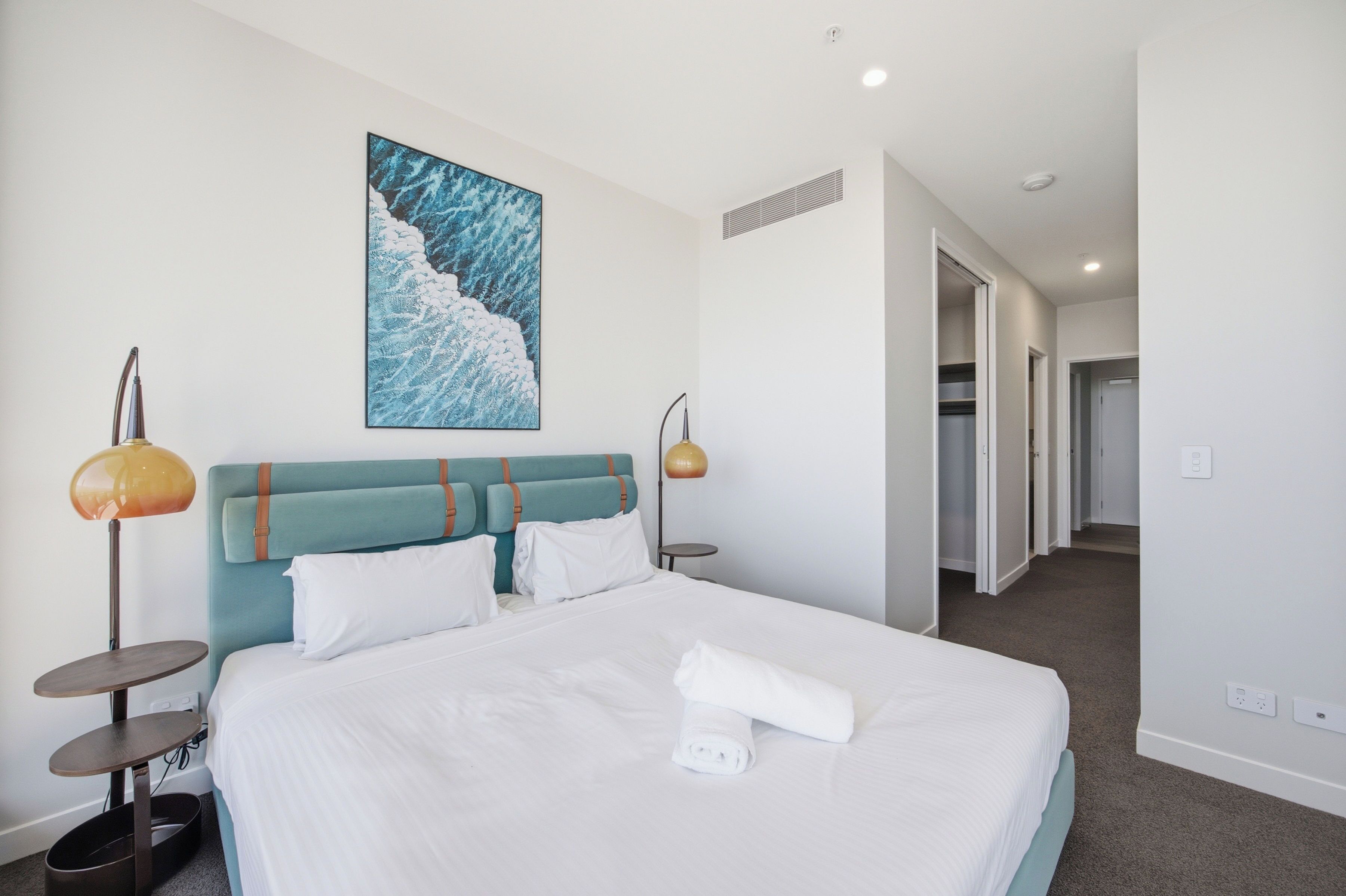 Habitación de hotel con una cama grande, cabecero, un cuadro, dos lámparas de noche y una cama bien hecha con una toalla enrollada.