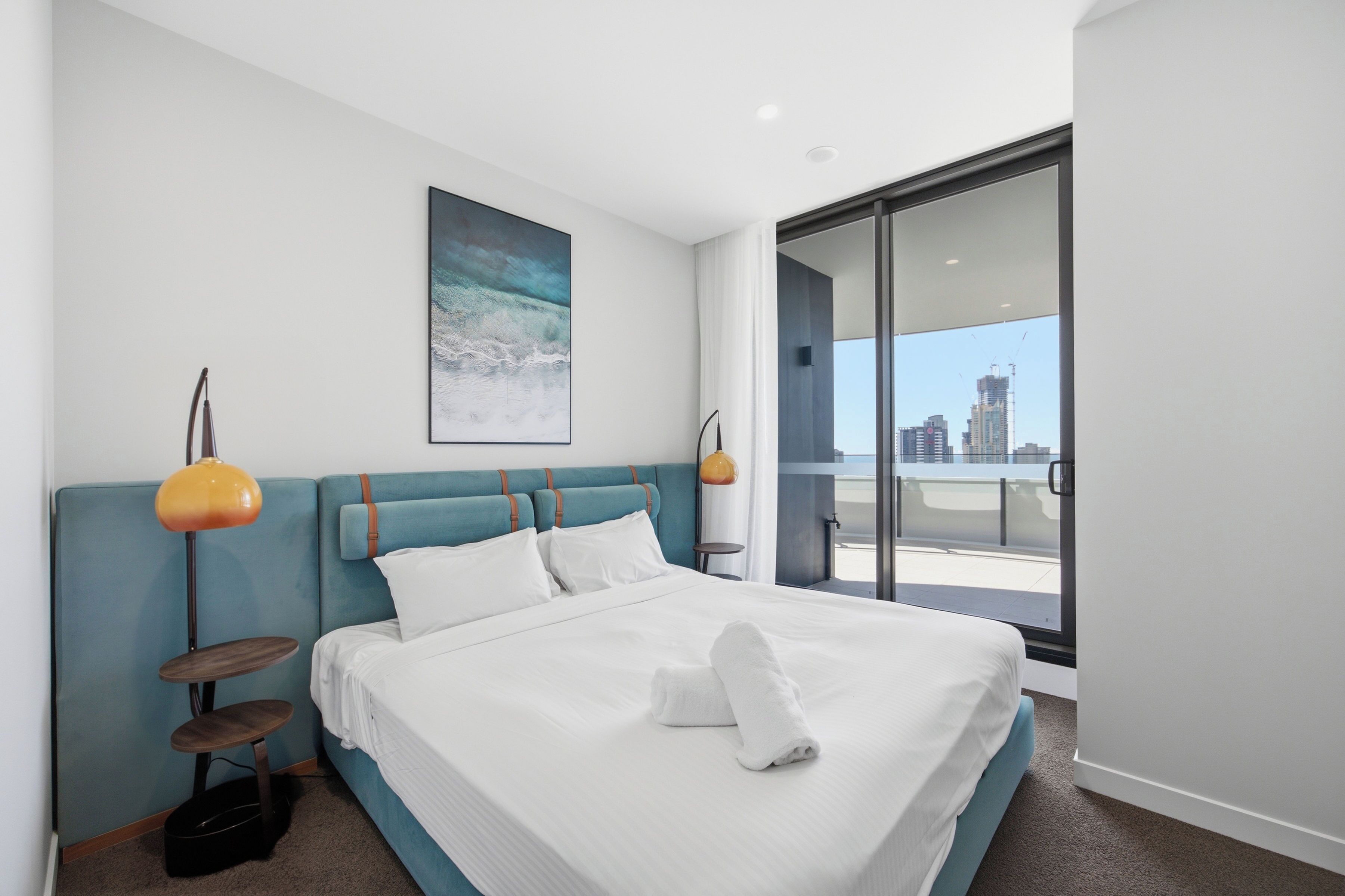 A modern hotel room with a large bed, two bedside lamps, a city view, and a painting on the wall.