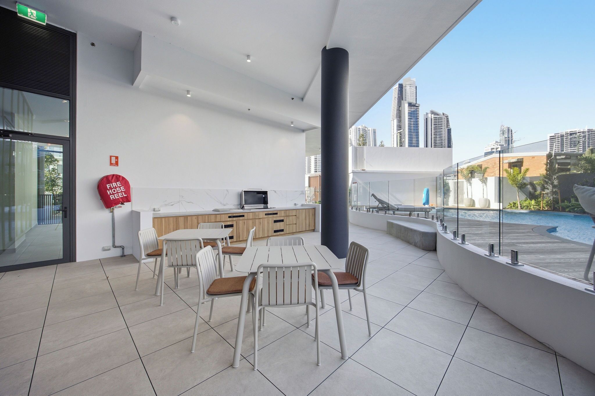 Un área de comedor moderna al aire libre con vista a una piscina, un carrete de manguera contra incendios y un paisaje urbano de fondo.