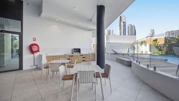 Un área de comedor moderna al aire libre con vista a una piscina, un carrete de manguera contra incendios y un paisaje urbano de fondo.