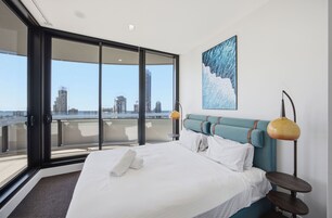 Quarto de hotel com uma cama, vista da cidade através de uma porta de vidro, um quadro na parede e um abajur.