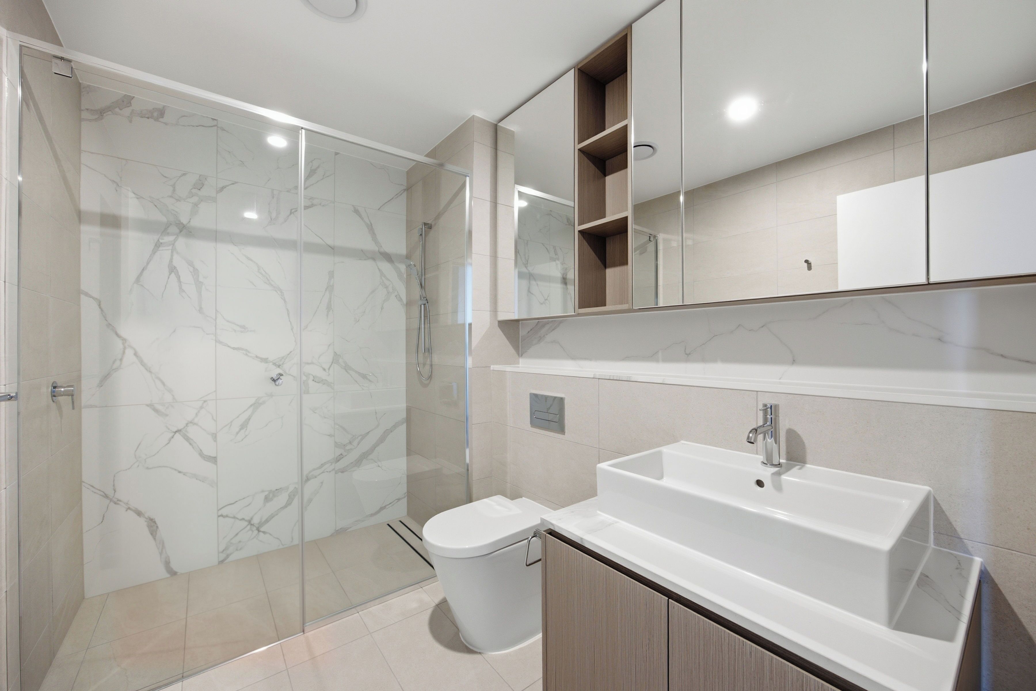 A modern bathroom with a glass shower enclosure, a white sink, and wooden cabinets.