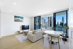 A modern living room with a city view, a flat-screen TV, a sofa, and a dining area with chairs and a table. - Chevron One Residences (Surfers Paradise)