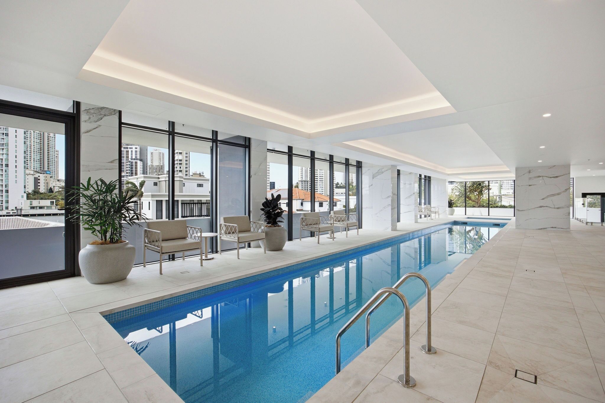 A modern indoor swimming pool with clear blue water, surrounded by lounge chairs and potted plants.