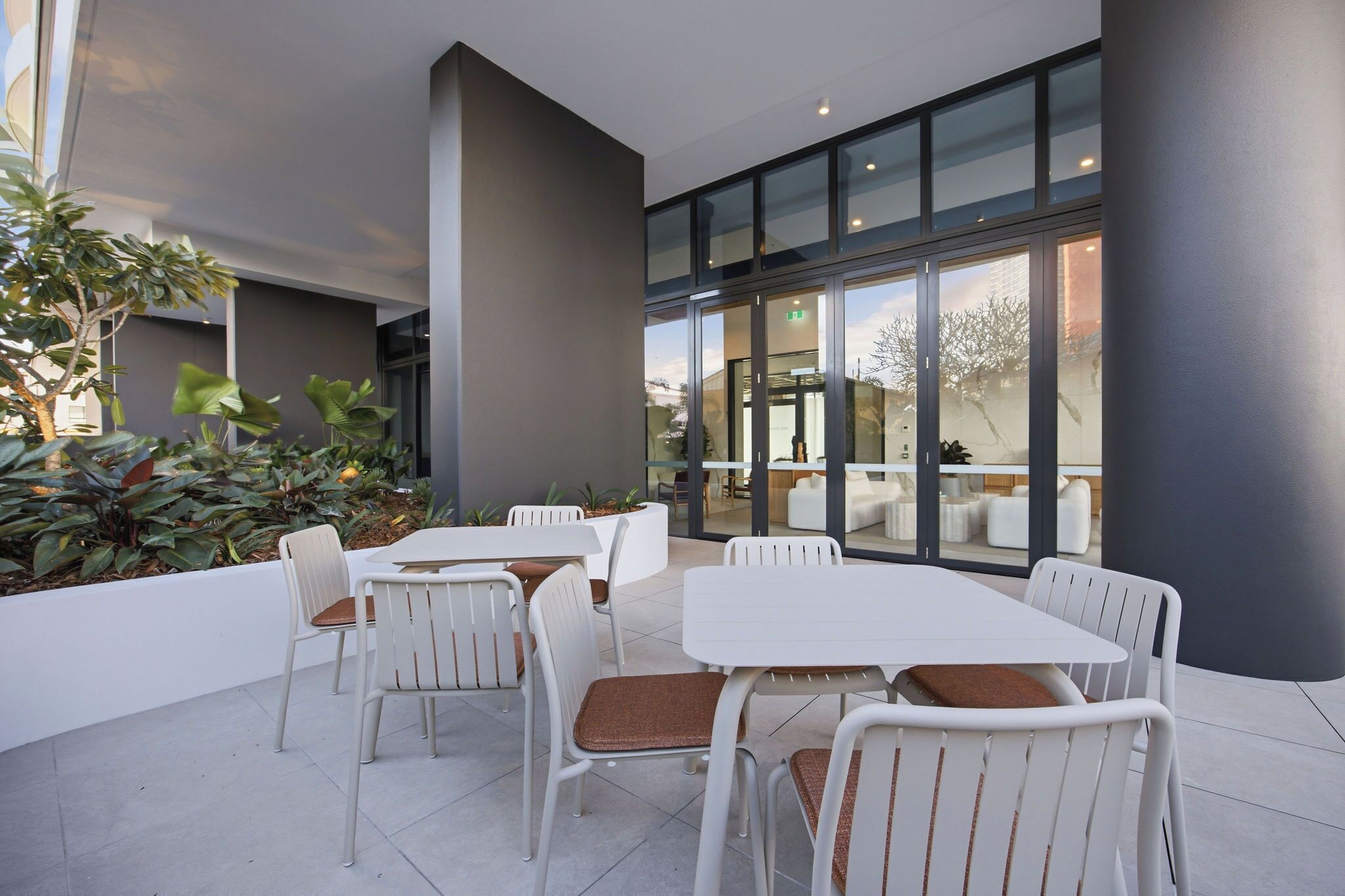An outdoor patio area with white tables and chairs, surrounded by plants and modern architecture.