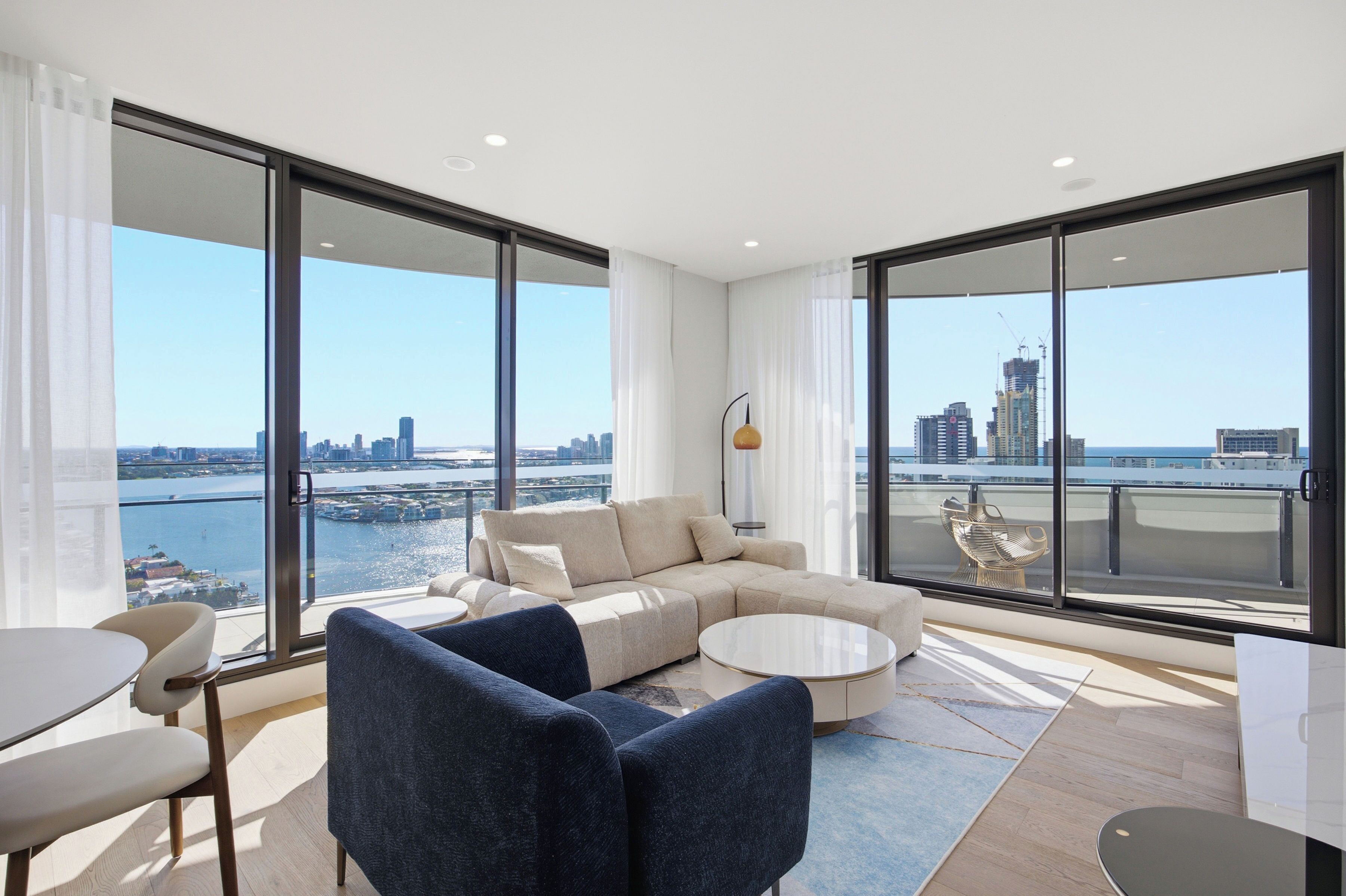 Una sala moderna con un sofá, sillas y una mesa de centro, con amplios ventanales que ofrecen vistas a la ciudad y al agua.