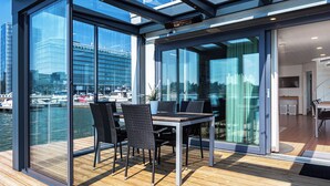 A modern room with a glass wall overlooking a marina, a wooden deck, and a dining area with chairs and a table.