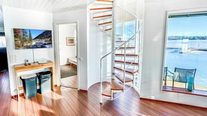 A modern interior with a spiral staircase, wooden flooring, a desk with a computer, and a large window overlooking a body of water.