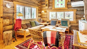 Ein rustikaler Innenraum einer Blockhütte mit einer Couch, Sesseln, einem Tisch und einem großen Fenster mit Buntglasdekor.