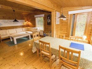 Puinen mökki, jonka sisustuksessa on sohva, ruokapöytä, tuoleja ja katosta roikkuva valaisin.