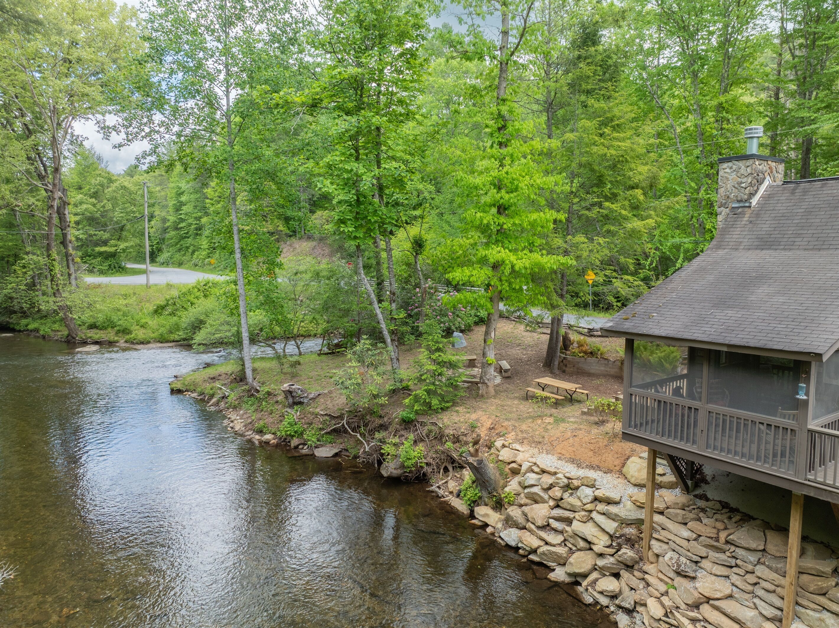 A cabin by a river with a stone wall, wooden deck, and a stone chimney.