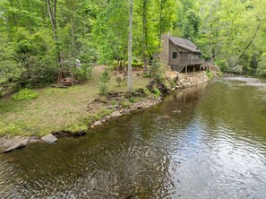 Eine Holzhütte am Fluss mit steinernen Fundamenten, umgeben von dichtem Wald.