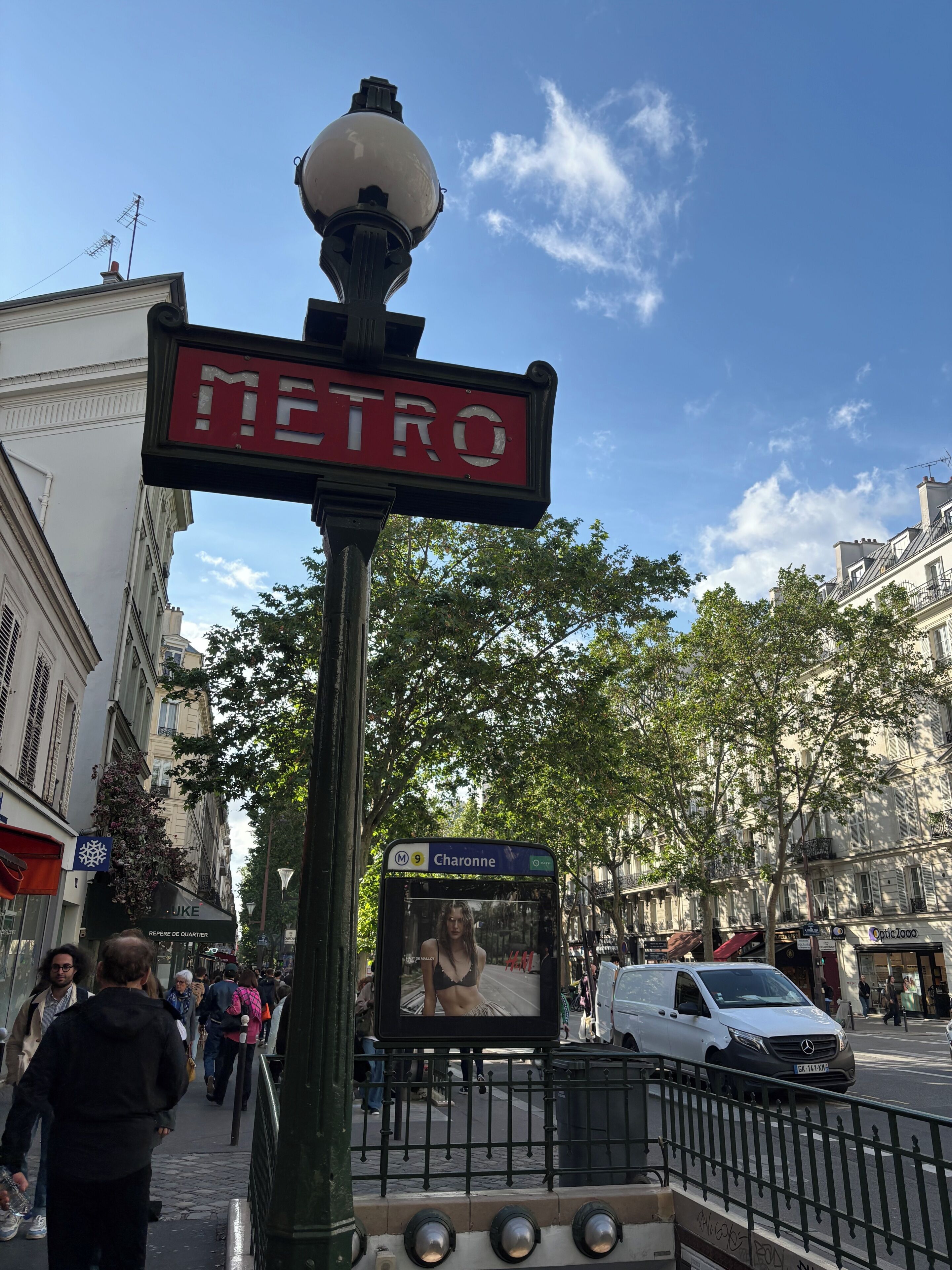 Une scène de rue avec un panneau indiquant une station de métro, un autobus affichant le numéro de ligne Charonne, et un fourgon blanc.