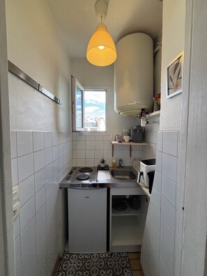 A compact kitchen with white tiles, a small refrigerator, a gas stove, a sink, and a window with a view of buildings. - Charming apartment in the heart of Paris (Île de France)