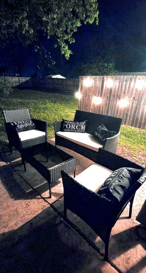 Un área de estar al aire libre con mobiliario de mimbre y una mesa de madera, iluminada con luces colgantes.
