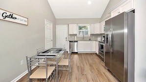 A modern kitchen with a stainless steel refrigerator, white cabinets, and a dining area with a glass table and metal chairs. - Bright 2BR/2BA Home. Large Backyard, 5 Gig Fiber. Nr. VBC, Arsenal, Space Center (Huntsville)