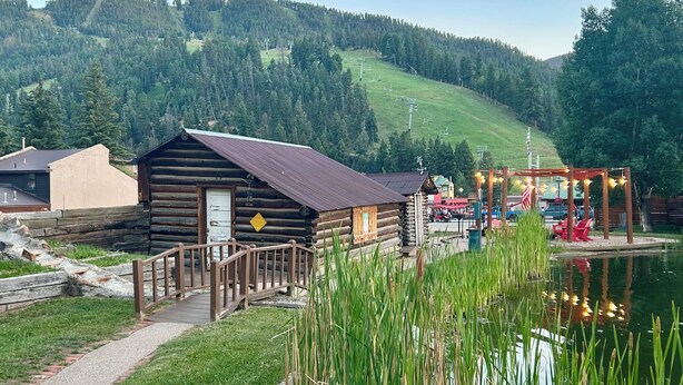 Un chalet rustique doté d’une terrasse en bois et d’un étang bordé de roseaux.