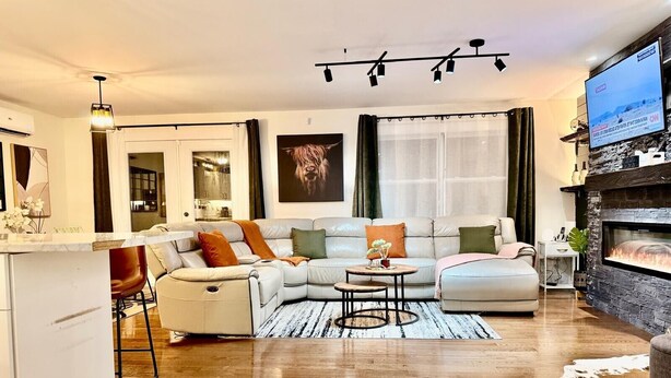 A modern living room with a sectional sofa, a fireplace, and a television mounted on the wall. - High-end haven where Serenity meets Style and Fun| 5mins to airport| 5 car slots (Moncton)