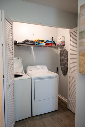 A laundry room with a washing machine, dryer, and a shelf with folded clothes. - Cozy Florida Retreat | Pool | Beach | Comfort (St. Augustine)