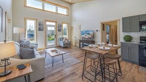 Amplia sala de estar con sofá, mesa de centro y mesa de comedor preparada para cuatro personas. Hay ventanas grandes, una cocina con electrodomésticos modernos y un televisor.