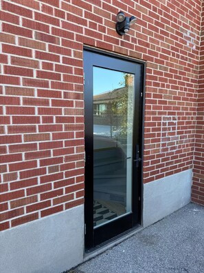 Un bâtiment en briques rouges, avec une porte noire et une caméra de surveillance au-dessus.