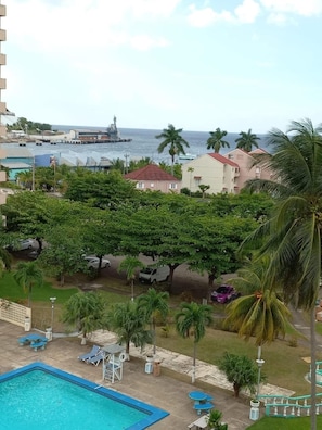 A pool area with lounge chairs, a clear view of the ocean, and a building with a red roof in the background. - Towers by the Beach is a super studio apartment that is centrally located. (Ocho Rios)