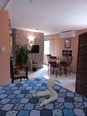 A living room with a sofa, a dining area with a table and chairs, a TV mounted on a stone wall, and a potted plant. - Towers by the Beach is a super studio apartment that is centrally located. (Ocho Rios)