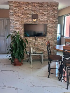 A living room with a stone wall, a flat-screen TV, a dining table with chairs, and a potted plant. - Towers by the Beach is a super studio apartment that is centrally located. (Ocho Rios)