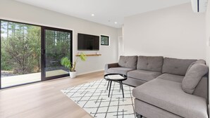 A modern living room with a grey sofa, a small black side table, a large window with a view of trees, and a mounted flat-screen TV. - Luxury Bush Retreat with Outdoor Bath, Birds, Trails & Wine Country  | Waimauku. (Helensville)
