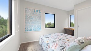 A bedroom with a large window, a bed with floral bedding, and a motivational quote on the wall. - Luxury Bush Retreat with Outdoor Bath, Birds, Trails & Wine Country  | Waimauku. (Helensville)