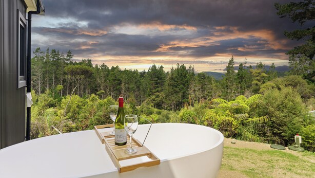 A round table with a bottle and glasses, overlooking a forested landscape with a cloudy sky. - Luxury Bush Retreat with Outdoor Bath, Birds, Trails & Wine Country  | Waimauku. (Helensville)