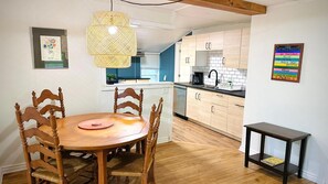 Un coin repas avec une table et des chaises en bois, un luminaire suspendu, une cuisine en arrière-plan et une petite table avec une chaise.