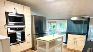 A modern kitchen with wooden cabinets, stainless steel appliances, a central island, and a window providing natural light. - Falcon Retreats  – Kestrel (Sharbot Lake)