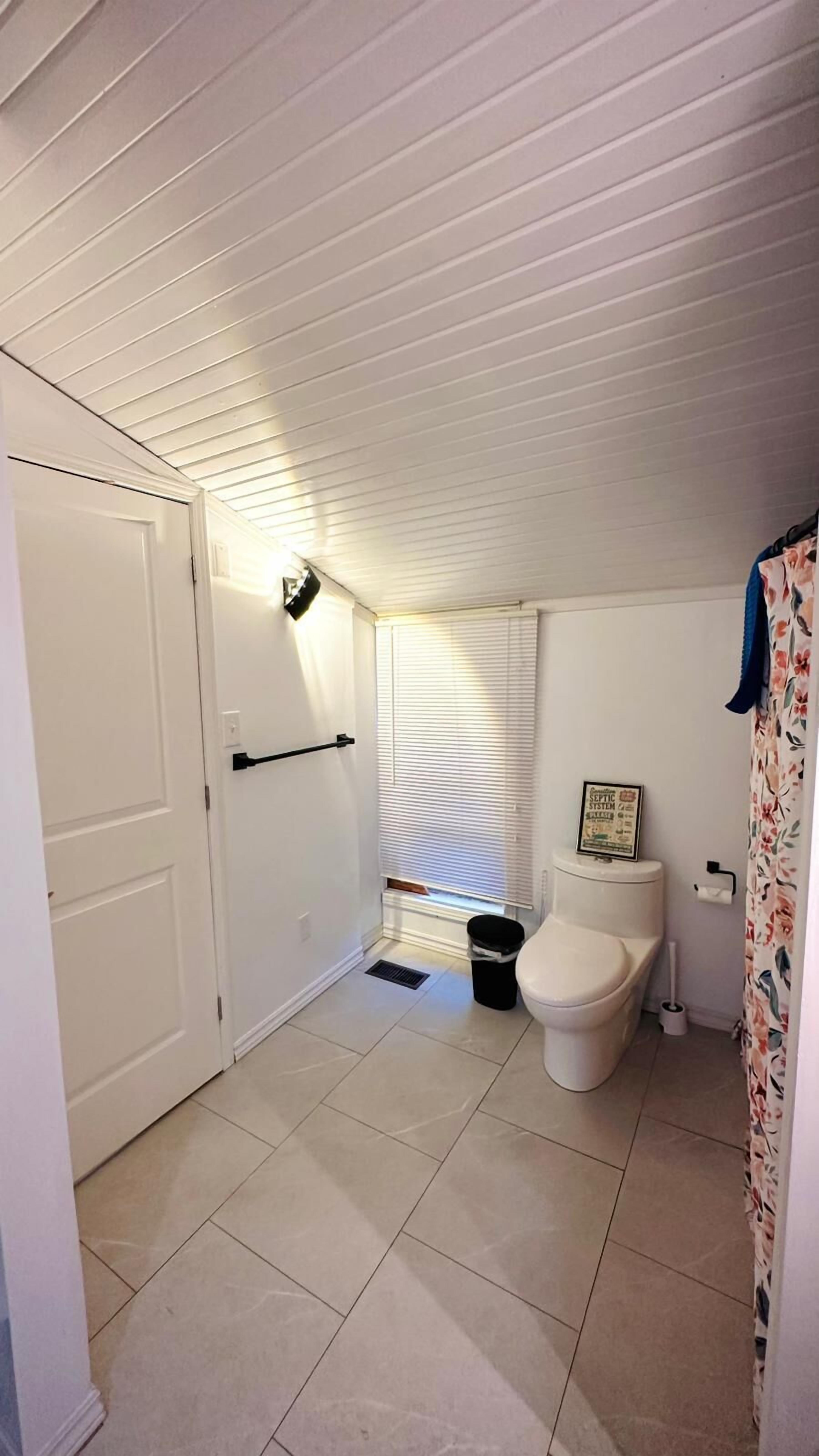 Un baño pequeño y limpio con suelo de baldosas blancas, un inodoro blanco, un cubo de basura negro y una ventana con persianas.