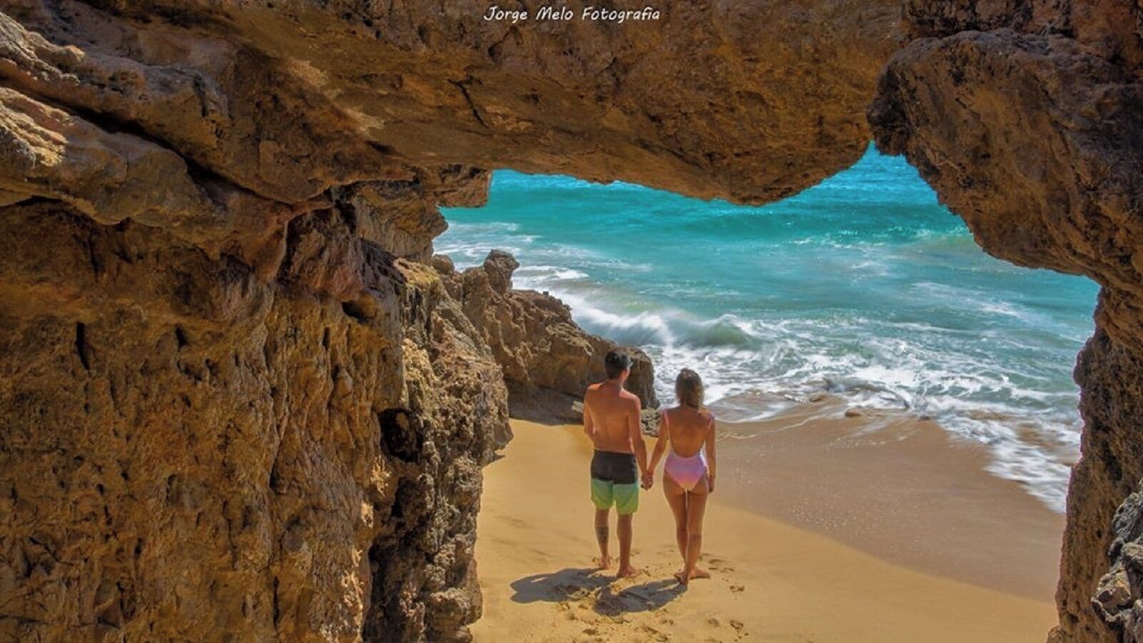 A couple standing at a beach through a rocky archway, overlooking the ocean.