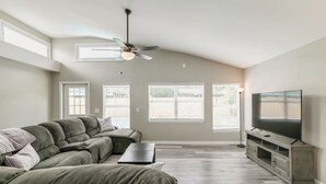 Una sala de estar con un sofá modular, un televisor de pantalla plana, un ventilador de techo y ventanas amplias.