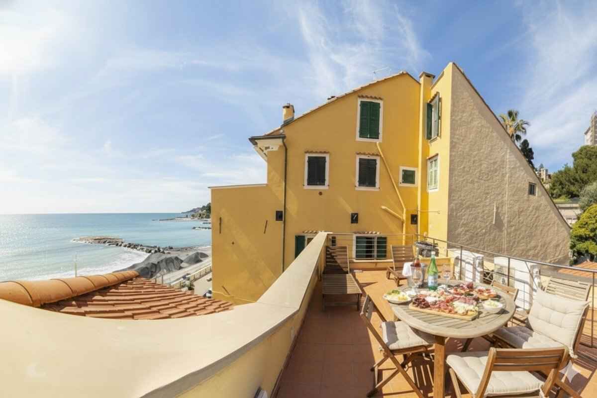 Un bâtiment jaune doté d’un balcon donnant sur la mer, une table avec de la nourriture et une chaise.